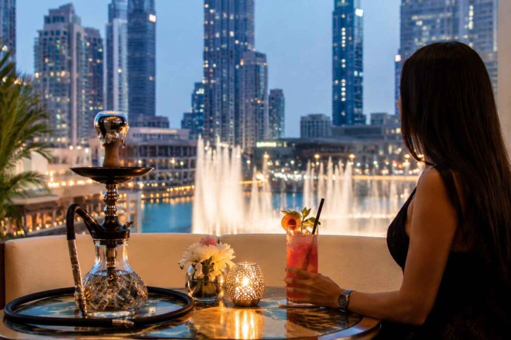 A woman seated at an upscale outdoor lounge overlooking the Dubai Fountain and downtown skyline at dusk, holding a pink fruit mocktail beside a premium glass shisha. A candle lantern and floral arrangement rest on the table, while illuminated skyscrapers and fountain displays create a sophisticated fine-dining atmosphere.