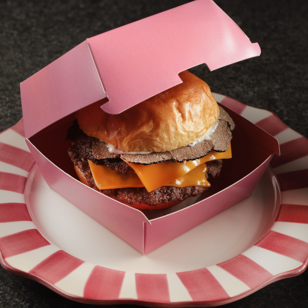Gourmet truffle cheeseburger served in a pink takeaway box on a striped plate.